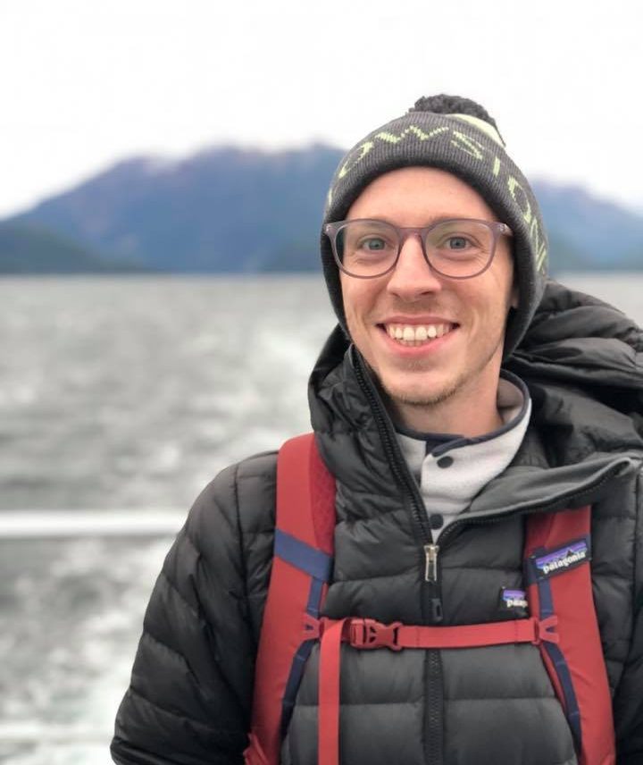 Head shot of a person wearing a puffy coat and a hat. A mountain is behind them, and they appear to be on a boat or ship on open water.