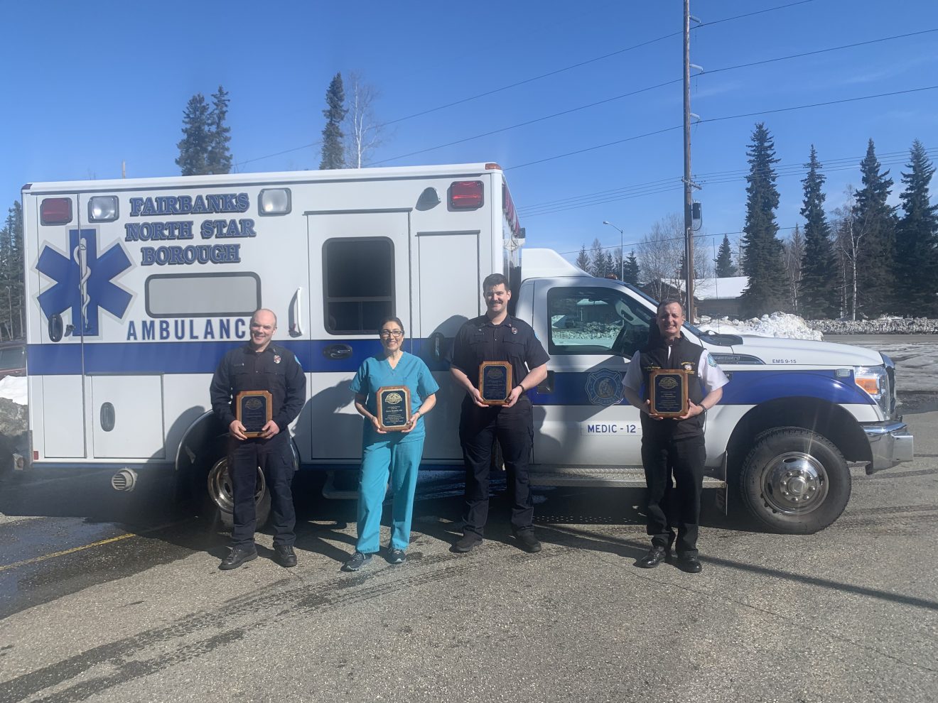 Three men and one woman wearing EMS or medical uniforms hold plaques while standing in front of an ambulance.