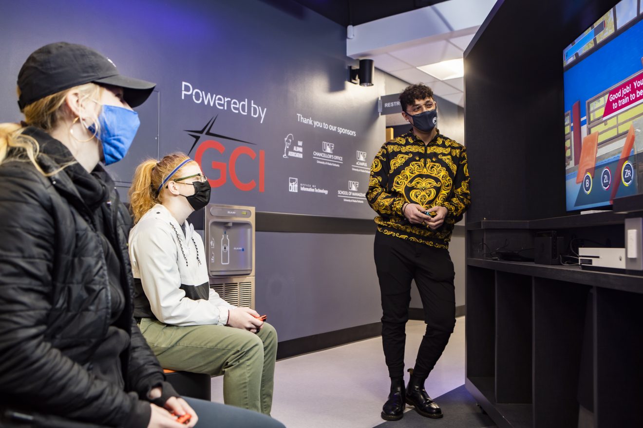Three students wearing masks sit or stand in front of a large gaming console.