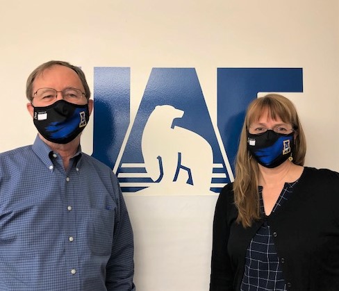A man and a woman, both wearing masks, stand in front of a wall with a UAF logo on it