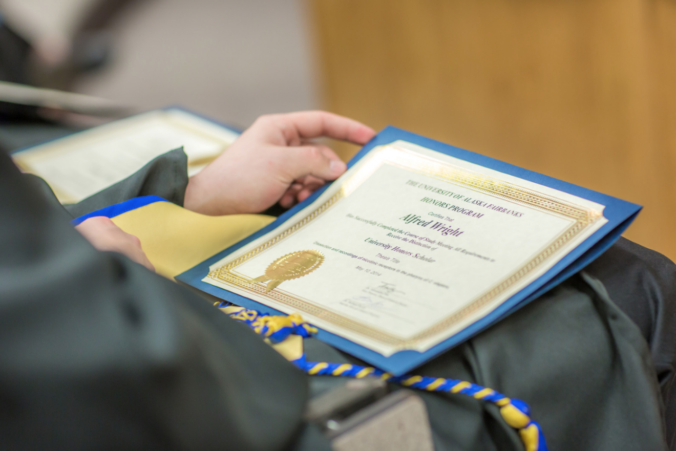 Closeup of diploma on the lap of someone wearing a robe and graduation cord