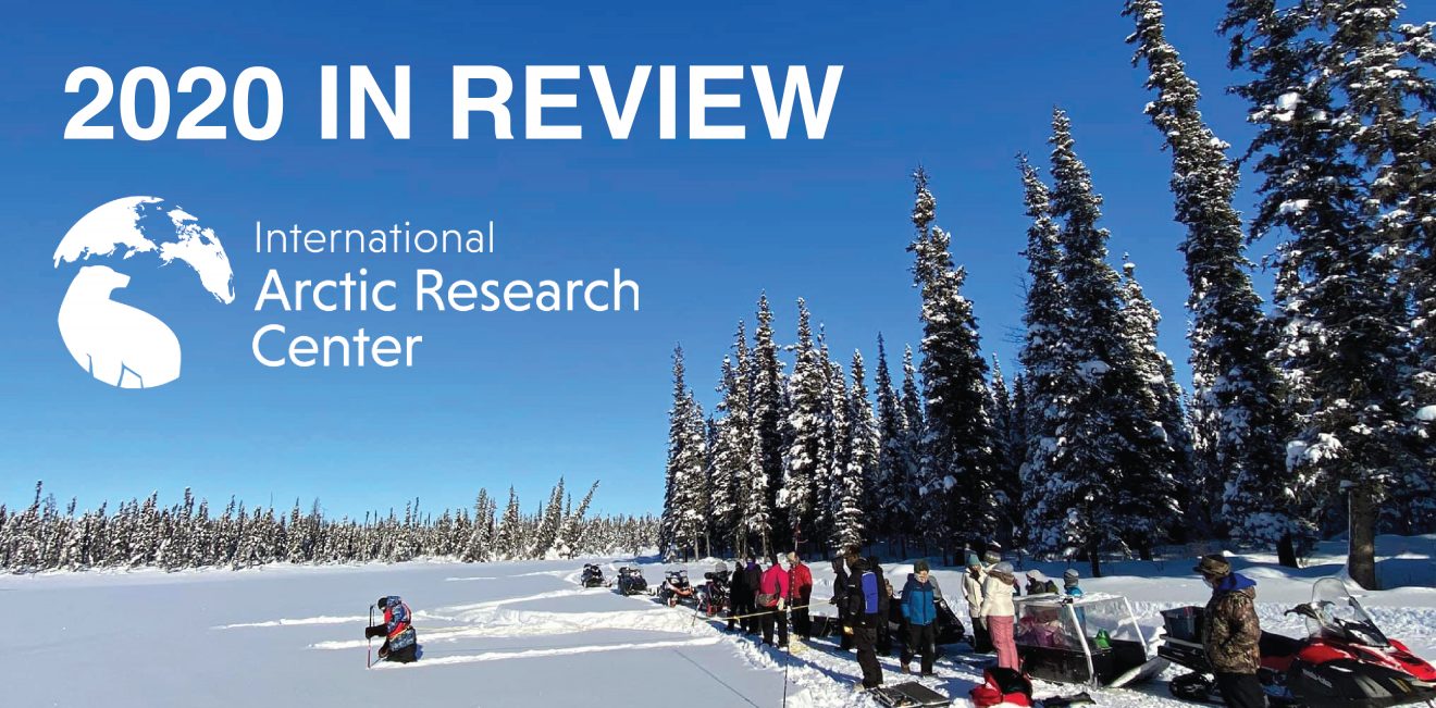 Large group of people standing on a frozen, snowy lake surrounded by trees. One person is farther out on the lake taking a measurement or sample.