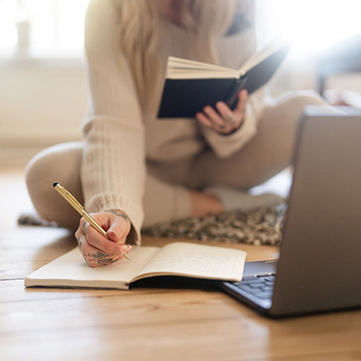 An anonymous person sits cross-legged on the floor in front of a computer, holding a book in one hand and taking notes with the other