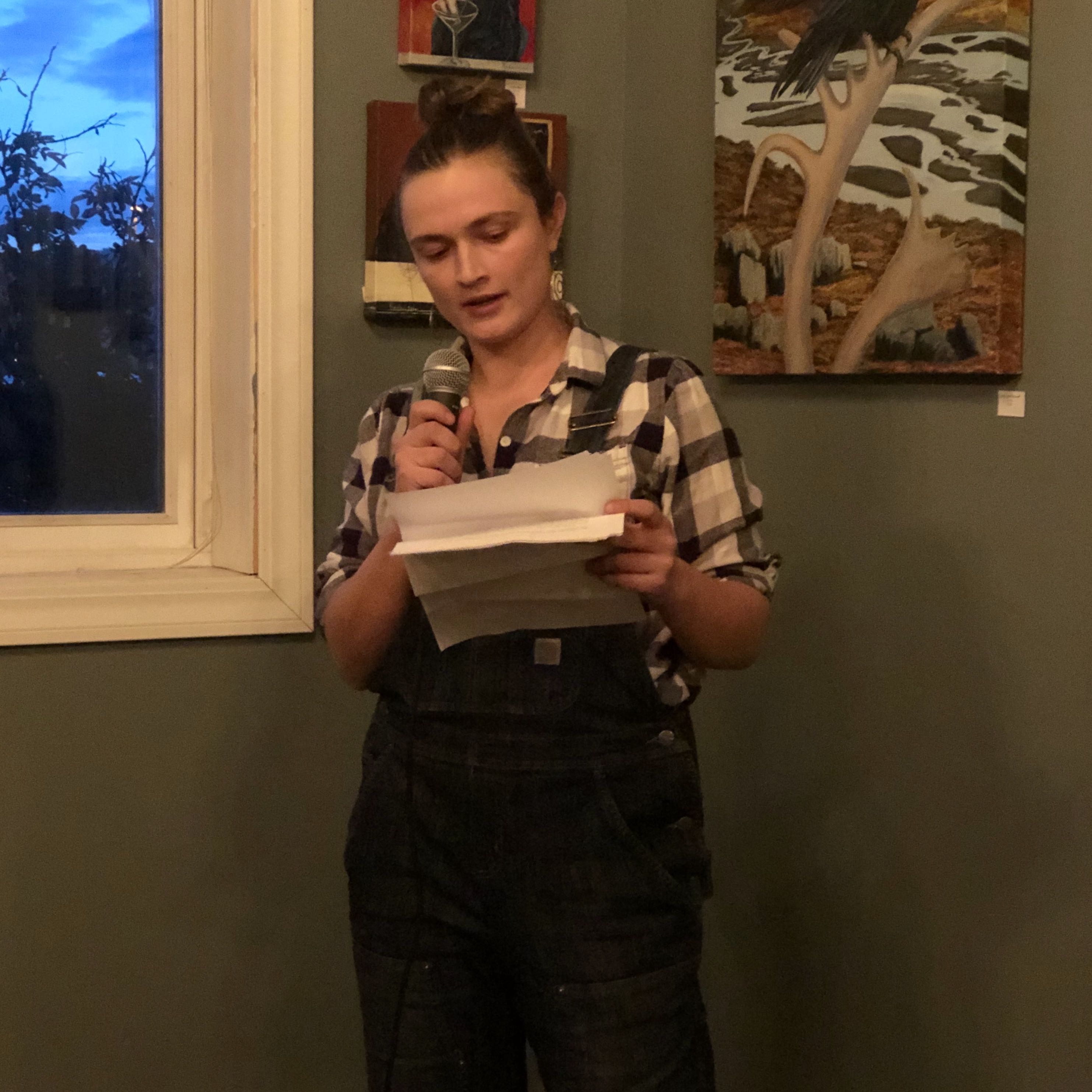 Keely O'Connell reads her nonfiction at an event in 2019. She wears denim overalls and a checkered shirt. She is holding a microphone.