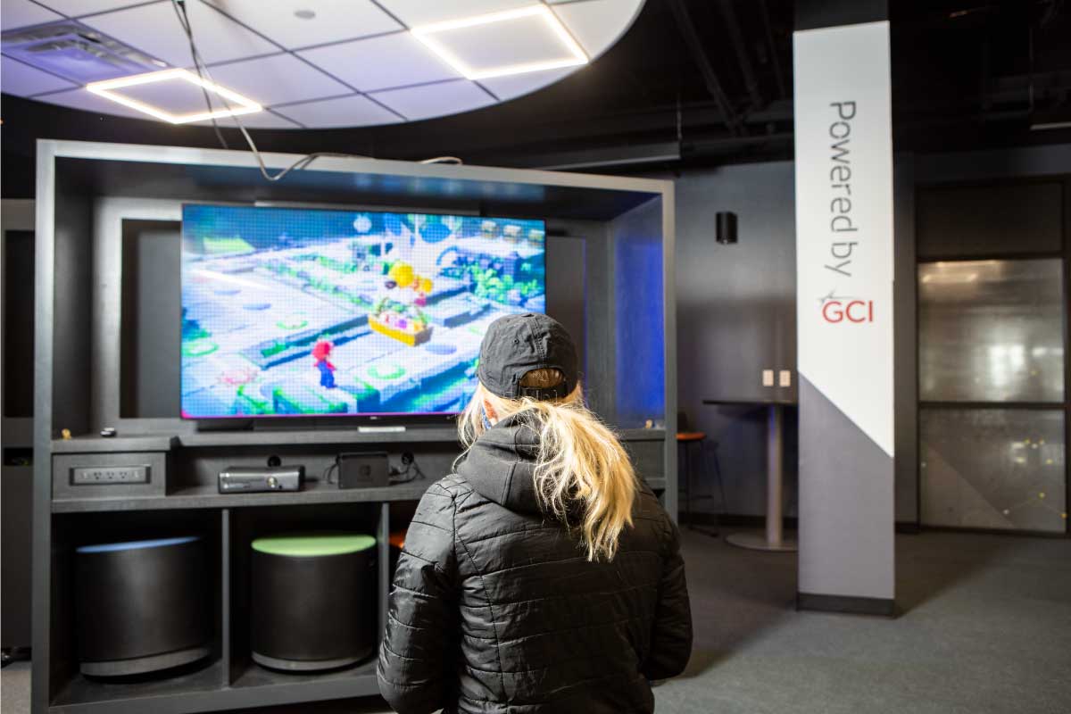 Student playing a game in the Alaska Esports Center console gaming area. A pillar with the GCI sponsor logo is in the background.