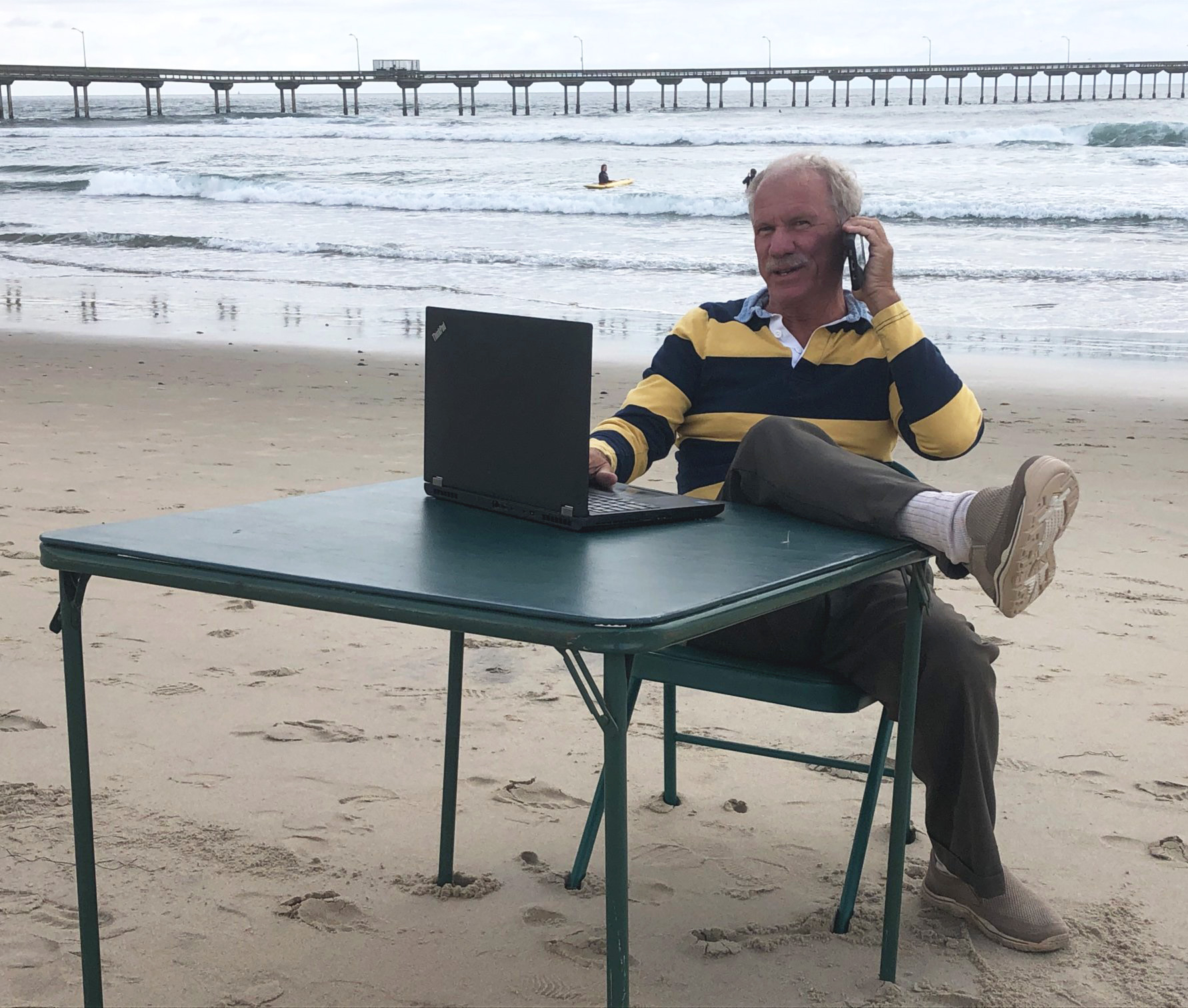A man sits at a card table on a beach while talking on the phone. A laptop computer is on the table. A pier extends into the ocean in the background.