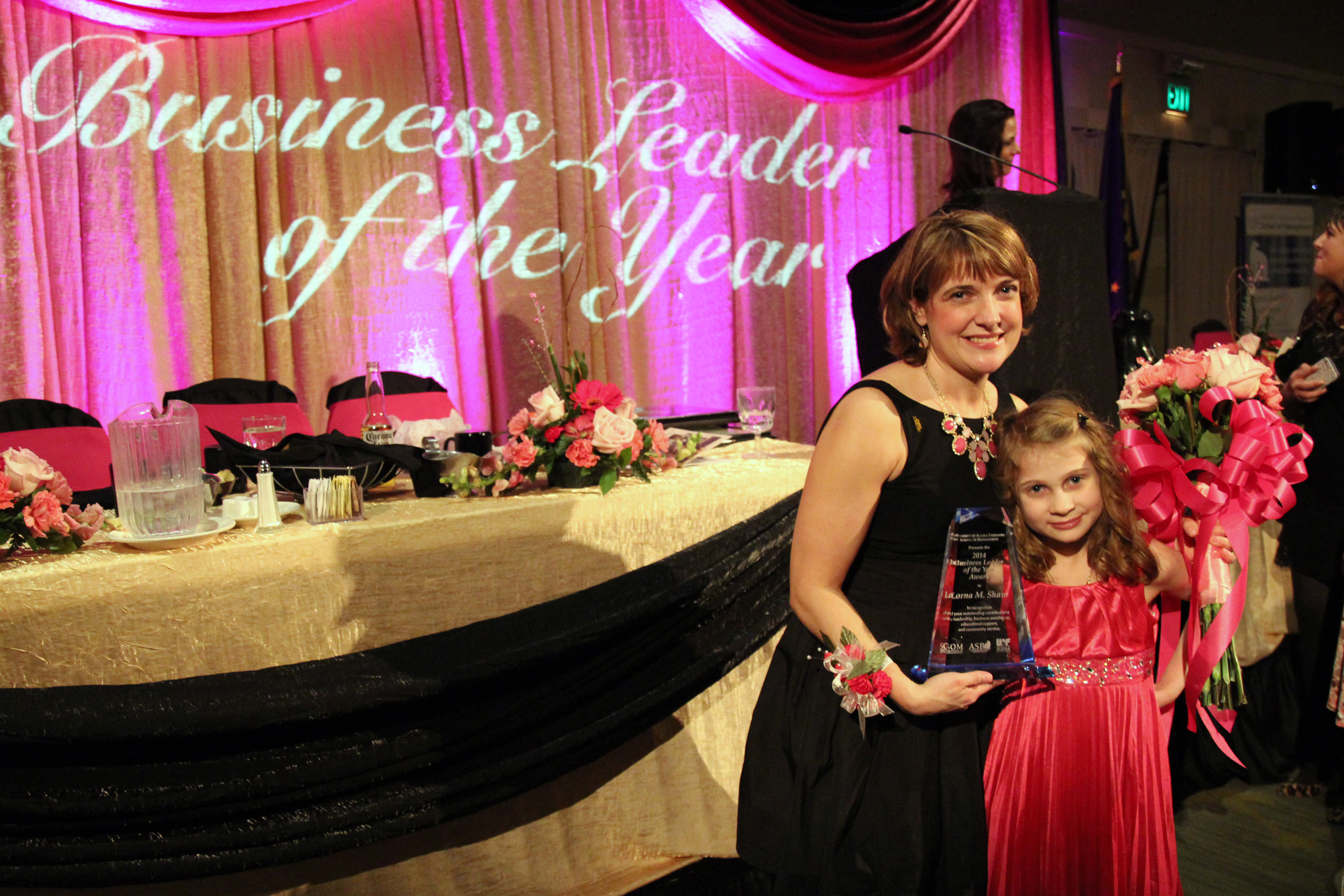 A woman in a dress stands before a stage table and holds an award, while embracing with one arm a young girl holding flowers. The words "Business Leader of the Year" are projected onto a pink stage curtain behind her.  