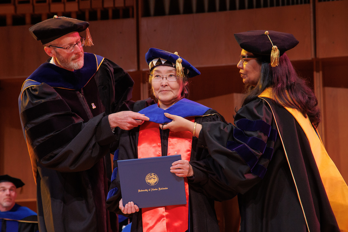Angass'aq Sally Samson stands while UAF Chancellor Dan White, left, and Provost Anupma Prakash perform the formal Ph.D.  hooding during ceremonies in the Davis Concert Hall on May 5. Samson earned her Ph.D. in Linguistics: Interdisciplinary Studies. Photo credit: Todd Paris