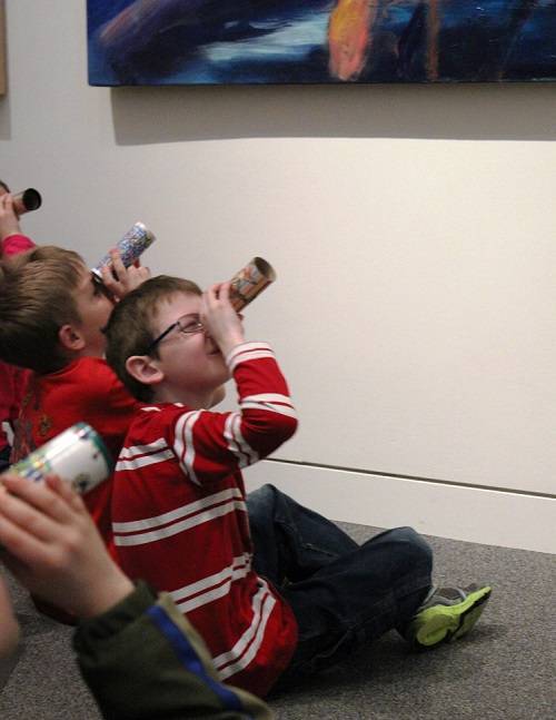 Children sitting on the floor, looking at an artwork through colorful cardboard tubes.