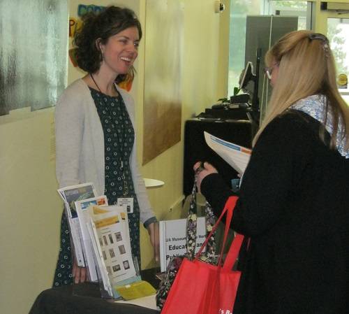 A museum employee talks to a teacher and shows her activity sheets, while they stand in the museum's main hallway.