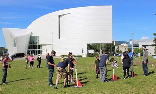 Several adults and children launching paper rockets on a large lawn. The museum building is in the background.