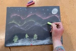 Child's hand holding a piece of green chalk and coloring a picture of the aurora.