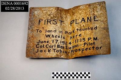 Rectangular sign made of rusted sheet metal, buckled inward in the middle. The sign is laying flat on a gray surface.