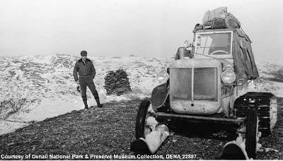 Black and white photograph of a man standing next to a M-7 snow jeep. The tractor is equipped with two wheels and two skis in the front. Behind each wheel is a rubber track.