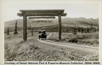 Black and white photograph of an automobile on a dirt road. There is a wooden arch over the road, with a hanging sign reading “Gateway to Mt. McKinley National Park".