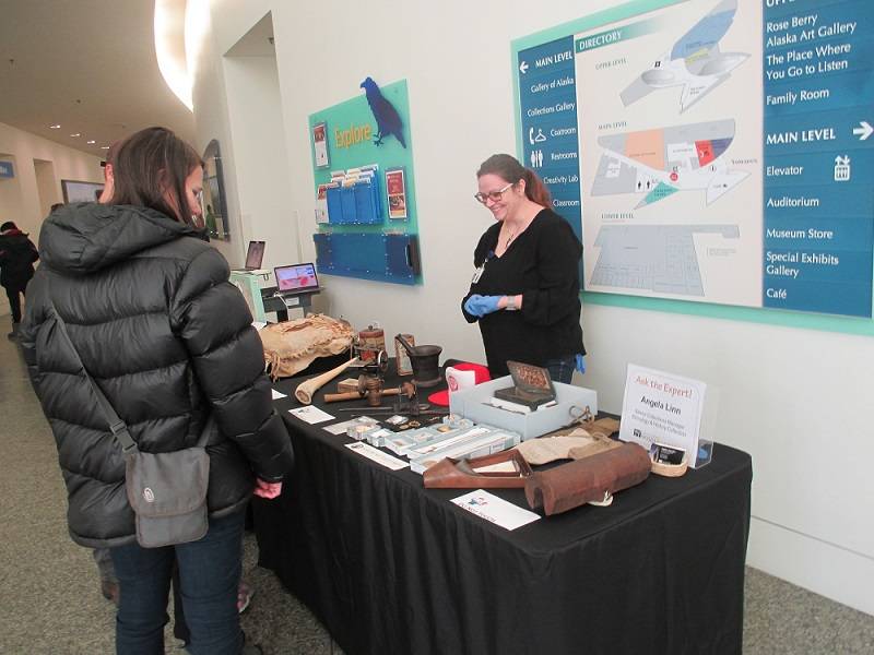 Person standing behind a table covered in a variety of museum objects. Two people are looking at the objects.