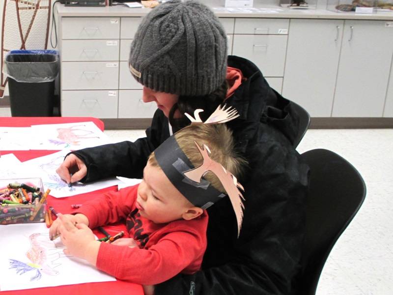 An adult and child use crayons to color pictures of moose. The child is wearing an antler headband made of paper.
