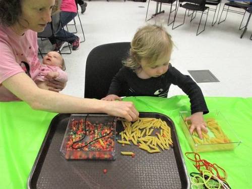 An adult holding a baby sits at a table with a small child, working on a craft project.