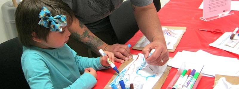 An adult stands behind a child sitting at a table. Both are using markers to color a model of a watershed.