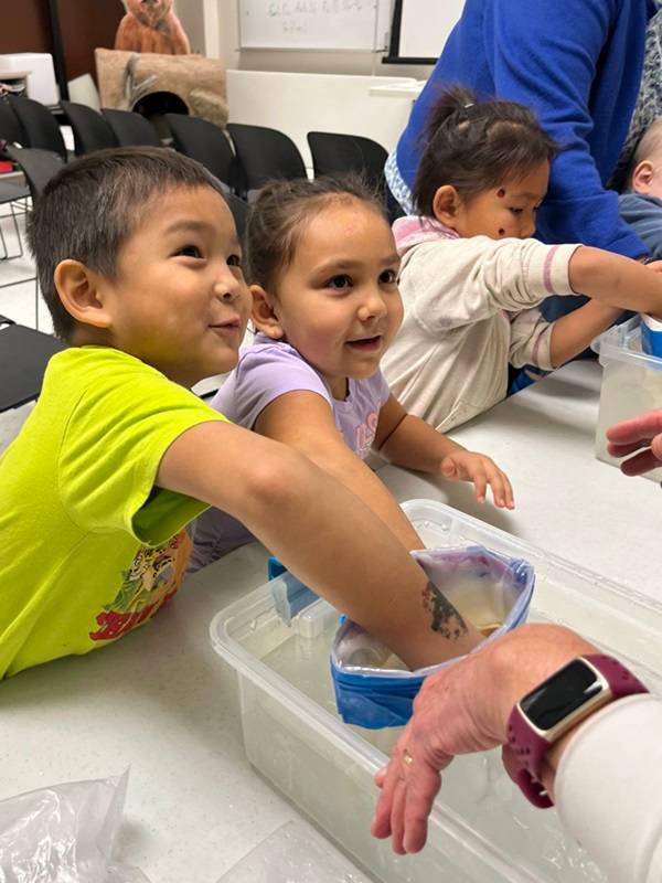 Two children smiling with their hands into Ziploc bags filled with shortening, dipping them into a tub of water.