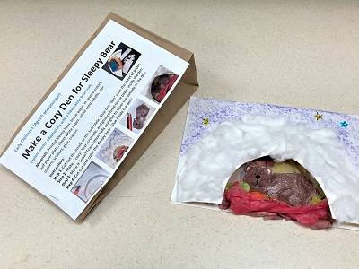 Winter Den craft example next to a brown paper lunch bag, with a instruction sheet labeled "Make a Cozy Den for Sleepy Bear" stapled to the bag.