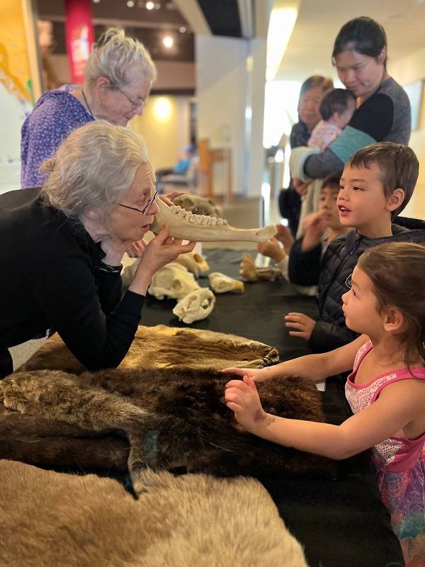 An adult stands behind a table holding an animal bone and talking to several children. Bones and furs are visible on the table.