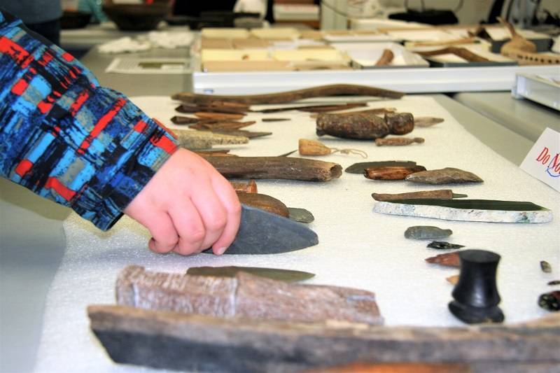 A variety of ancient tools on a table. A hand is reaching out to touch one of them.