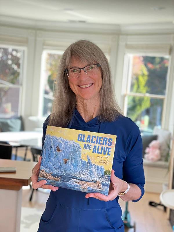 Debbie Miller, an older woman with long gray hair and a blue shirt, holds a copy of the book "Glaciers Are Alive".