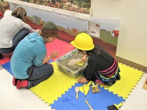 Adults and children dig for fossils in a sandbox.
