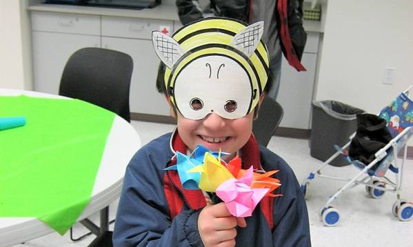 Child wearing a paper bumblebee mask and holding origami flowers.