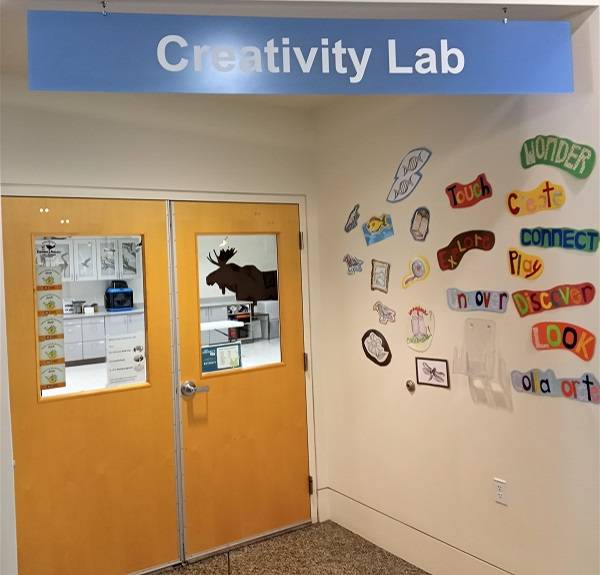 A sign saying "Creativity Lab" hanging  over a set of double doors. A variety of brightly colored pictures are on the wall next to the doors.