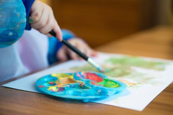 A child's hands holding a paintbrush next to a plate with several colors of paint.