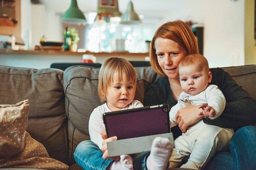An adult sits on a couch with two young children, looking at a computer tablet.