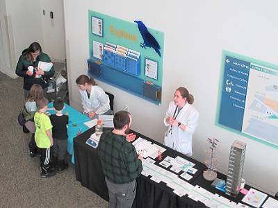 Two scientists stand behind a table with a DNA timeline, DNA model, and various pipettes and other lab equipments. Several kids and adults are looking at the display.