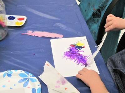 Closeup of a child's hand using a feather to paint a piece of paper. The paper has purple paint on it. 