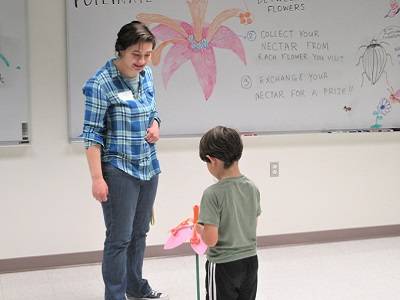 Child placing pom poms in a foam flower model, as an adult looks on. Illustrations of flowers are visible on a whiteboard.