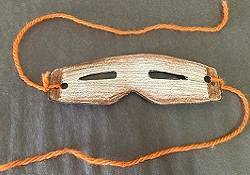 Snow goggles made of paper and colored to look like wood. Pieces of orange yarn are tied to each side.
