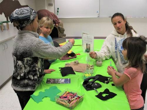 Children sit and stand around a table, working on craft projects.