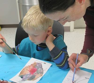 A child holds a paintbrush over a watercolor painting of a grass basket. An adult also holding a paintbrush leans over the child.