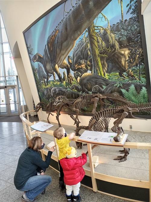 An adult and two children look at dinosaur skeletons and a dinosaur mural in a museum lobby.