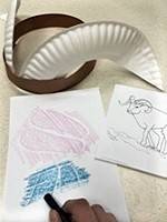 A Dall sheep headband made with a paper plate and a strip of brown paper, next to a drawing of a Dall sheep. A crayon track rubbing is next to the drawing.