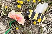 A bee made from construction paper and colorful paper flower, sitting on the ground next to dandelions.