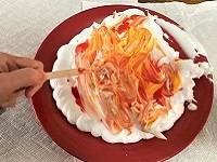 Hand using a popsicle stick to sworl red and orange food coloring into shaving cream.