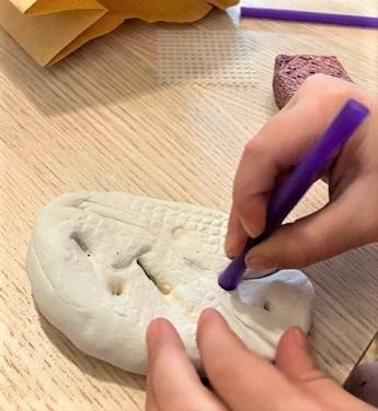 Child's hands holding a purple straw and making designs on a piece of white clay.