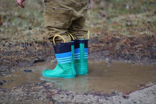 Children wearing teal rain boots stepping in a mud puddle.