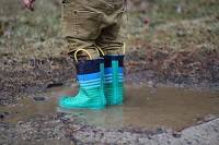 Child wearing teal rain boots stepping in a mud puddle.