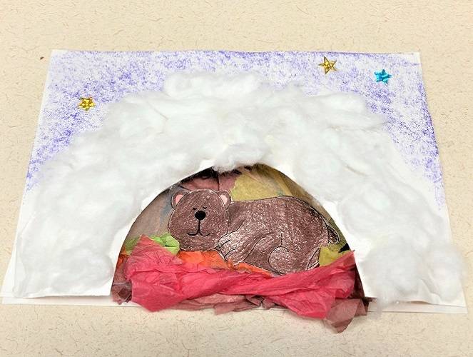 Half of a paper plate with the inside cut out, glued to a sheet of paper, decorated with pieces of colorful tissue paper, cotton balls, and star stickers. A drawing of a brown bear is in the den.