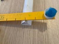 Yellow stick rested on a fulcrum made from a folded piece of paper. A blue pom-pom rests on one end of the stick. The stick has black lines marking one inch intervals.