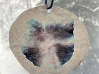 Circle of coffee filter paper with a bluish-black design drawn on it. The colors are faded at the edges. A piece of blue yarn is tied through one edge.