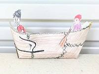 Model umiak made out of paper and decorated to look like walrus hide. Cutouts of two people are glued inside the boat.